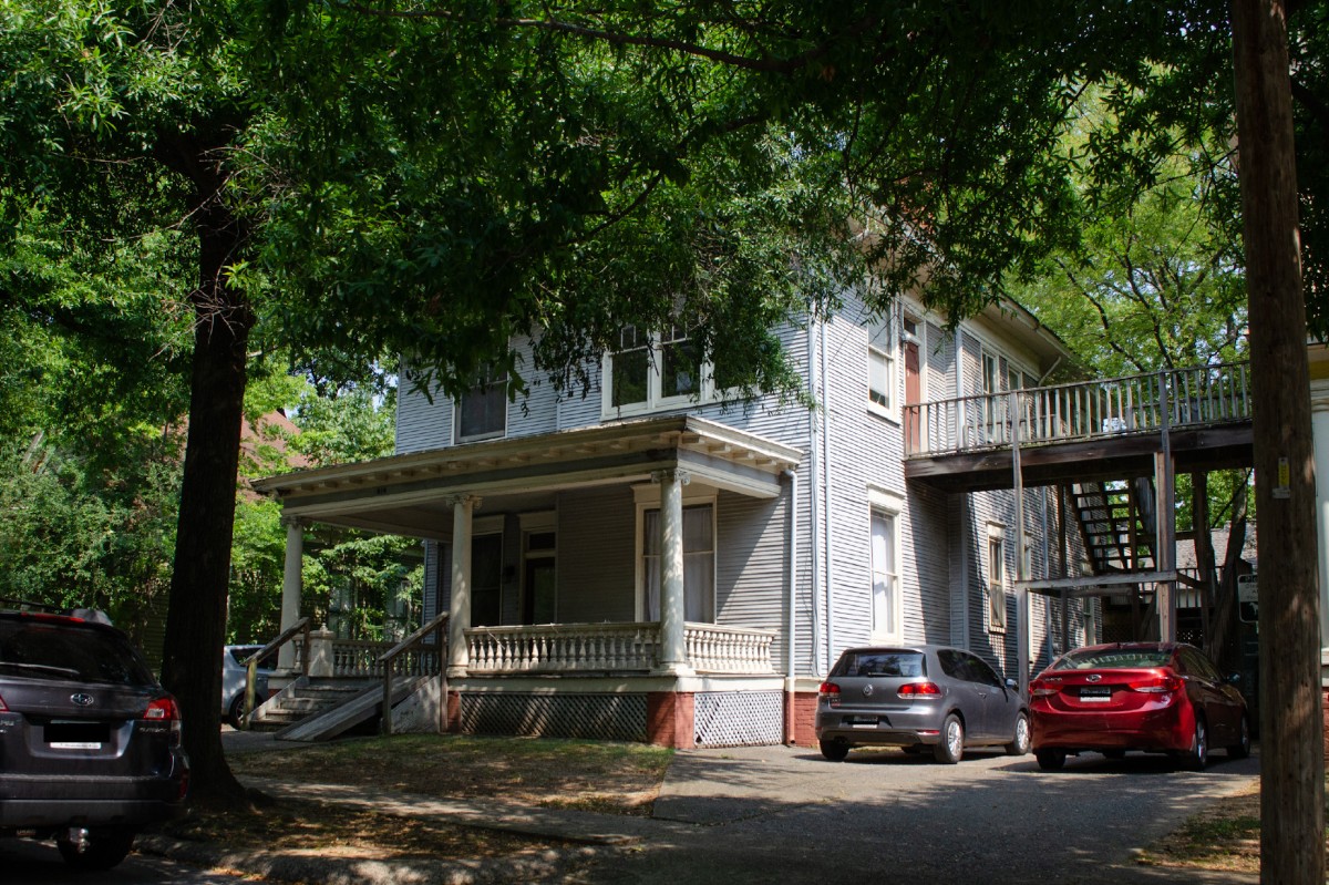An old two-story, gray-blue house with a porch on a tree-lined street. The scene is peaceful. Several cars are parked on the side of the house.