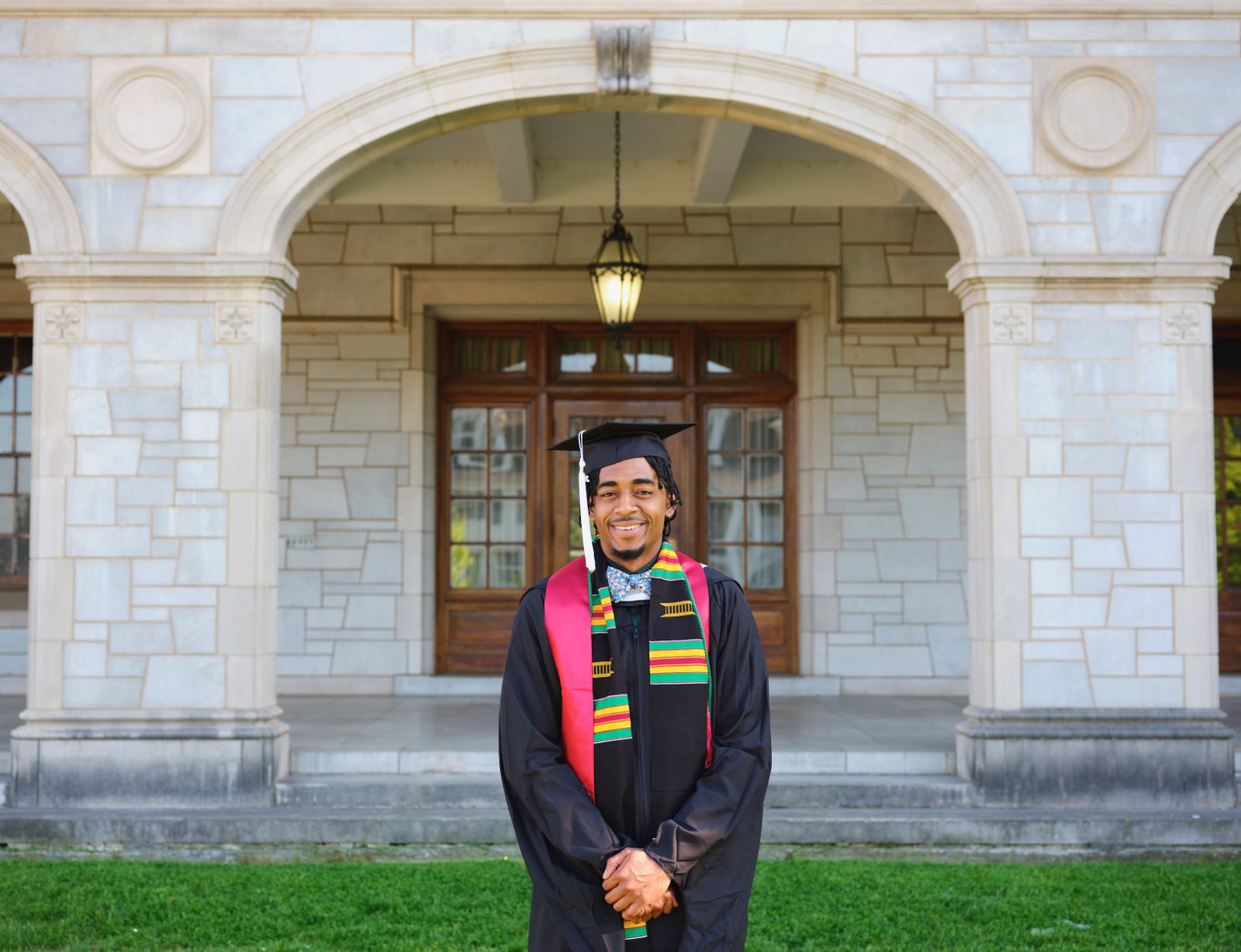 A young black man in a graduation cap and gown standing in front of a building with a marble facade